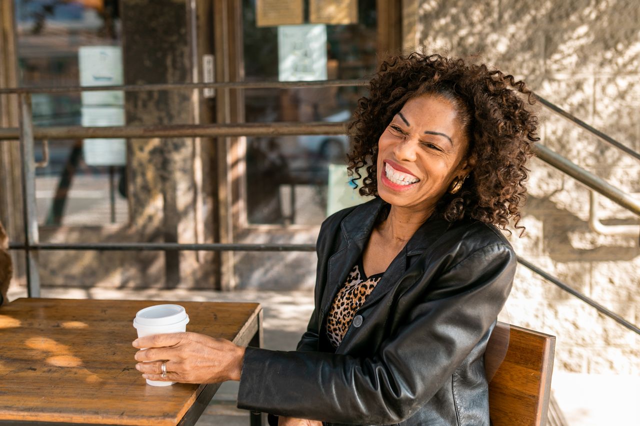 home-hero-02 Smiling senior woman relaxing with a coffee at an outdoor cafe, embodying elegant senior style.